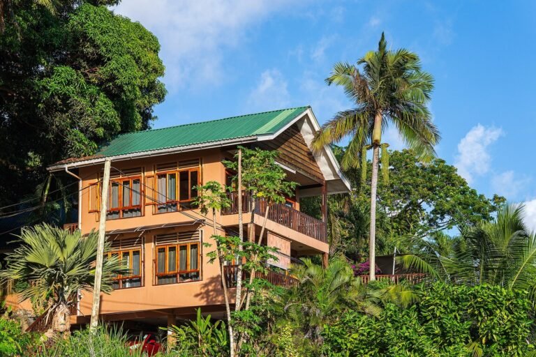 一栋两层住宅楼房坐落在绿树环绕的山坡上,橙色外墙配绿色金属屋顶,木框窗户与阳台清晰可见,四周棕榈树与热带植被茂盛生长,蓝天白云为背景